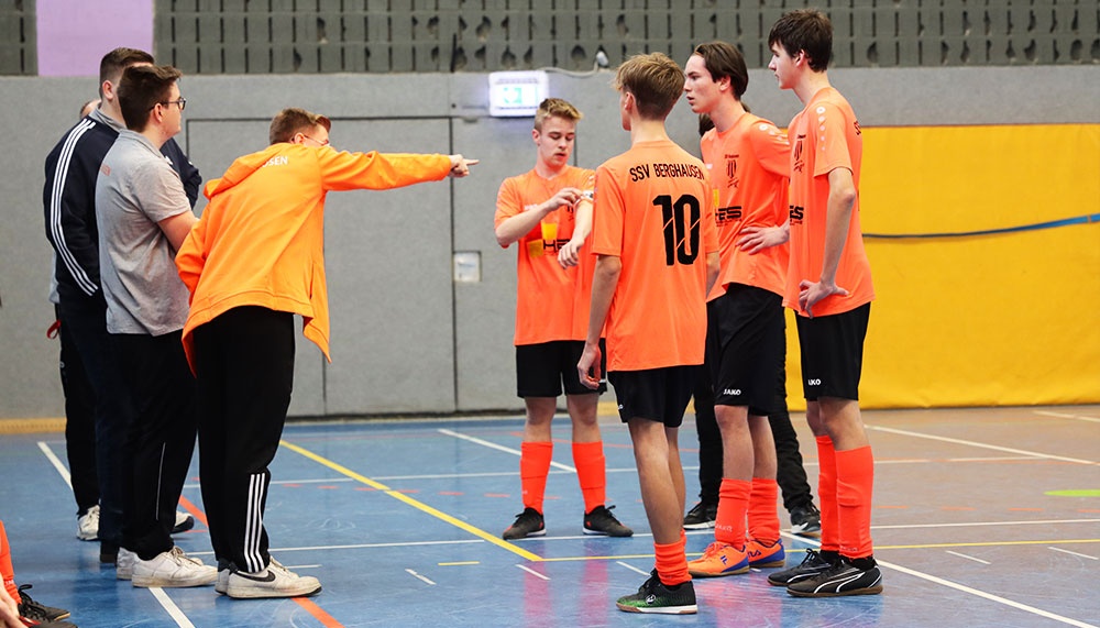 [{"clang_id":1,"value":"Handballteam in orangefarbenen Trikots und Jacken in Sporthalle. Trainer erklärt Spielstrategie, Spieler hören aufmerksam zu."}]