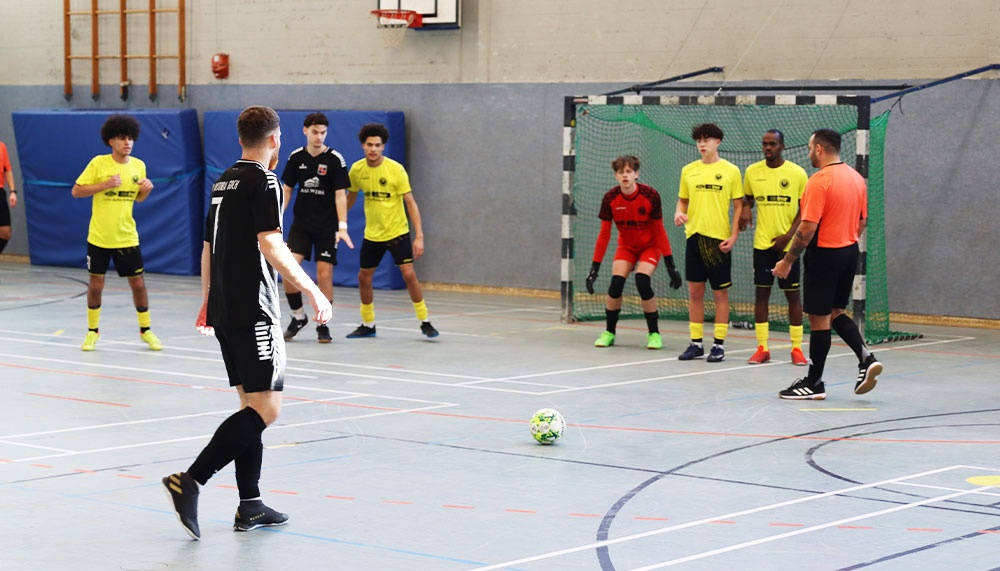 [{"clang_id":1,"value":"Futsalspiel in Sporthalle. Ein Spieler schießt auf das Tor, während andere Spieler zuschauen. Tragen Sportkleidung in Schwarz, Gelb und Orange."}]
