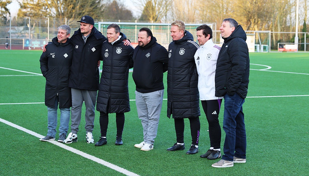 [{"clang_id":1,"value":"Gruppe von acht Männern auf einem Fußballfeld. Sie tragen überwiegend dunkle Jacken und Trainingskleidung, einige mit Vereinslogos."}]