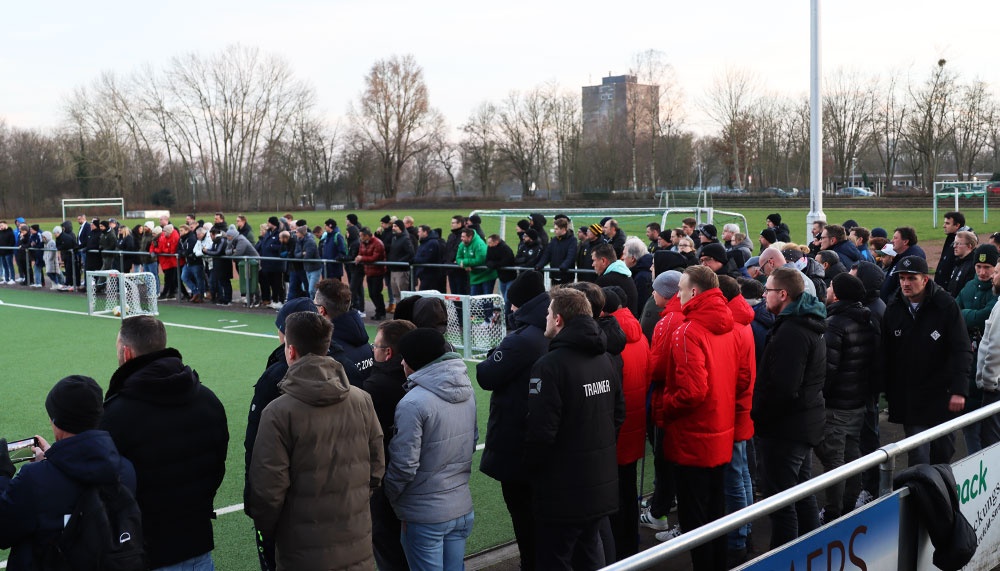 [{"clang_id":1,"value":"Zuschauergruppe am Spielfeldrand eines Fußballplatzes. Viele Menschen stehen dicht gedrängt, einige tragen Winterjacken. Im Hintergrund sind Bäume und der Platz erkennbar."}]