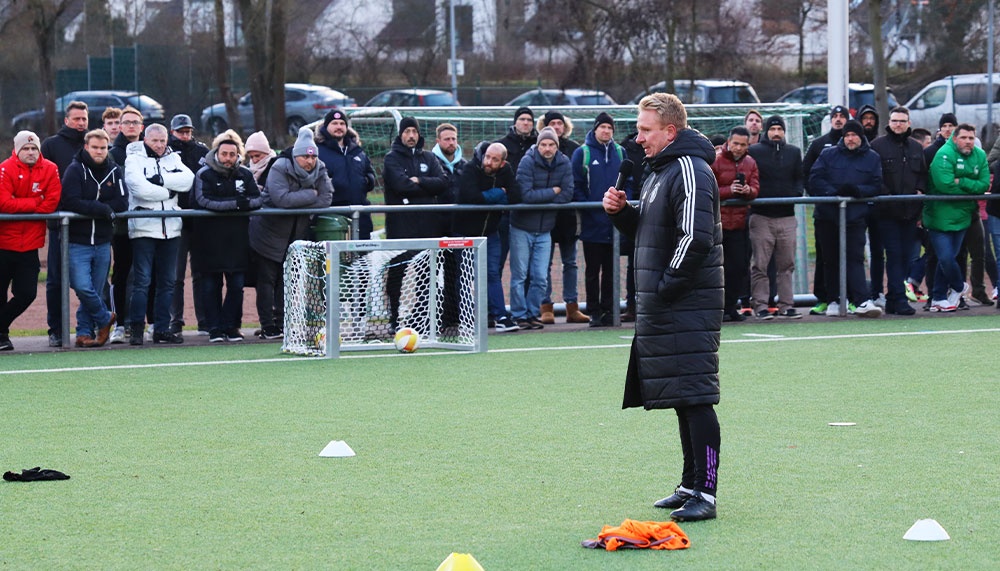 [{"clang_id":1,"value":"Fußballfeld mit Trainer, der vor einer Gruppe von Männern spricht. Im Hintergrund ein Fußballtor und Bäume."}]