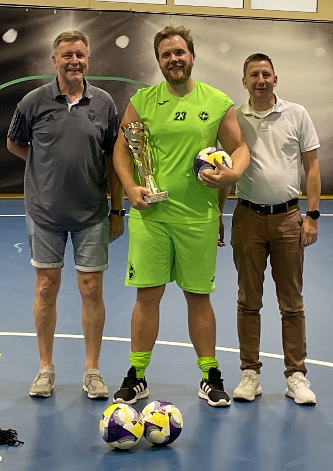 [{"clang_id":1,"value":"Drei Männer stehen in einer Sporthalle. Der Mittlere hält einen Pokal und eine einen Fußball. Alle tragen Sportkleidung. Vor ihnen liegen zwei Fußbälle."}]