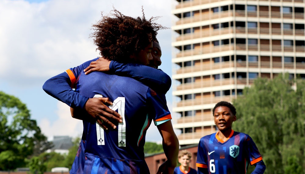 [{"clang_id":1,"value":"Zwei Fußballspieler im blauen Trikot umarmen sich vor einem hohen Gebäude und einer Grünfläche. Im Hintergrund weitere Spieler."}]