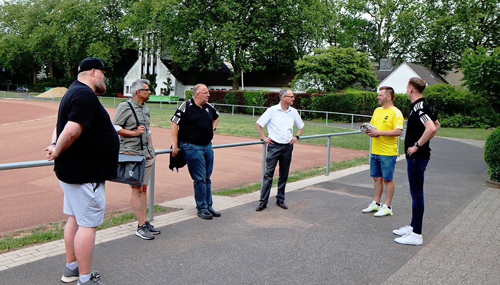 [{"clang_id":1,"value":"Sechs Männer stehen und sprechen neben einem Sportplatz. Im Hintergrund Häuser und Bäume. Einige tragen Sportkleidung, andere Alltagskleidung."}]