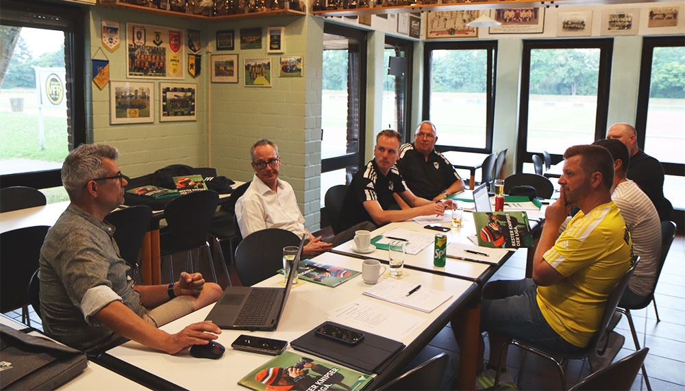 [{"clang_id":1,"value":"Sitzung in einem Raum mit Blick auf einen Sportplatz. Sechs Männer sitzen um einen Tisch, besprechen Unterlagen und schauen auf einen Laptop. Wände mit Fotos."}]