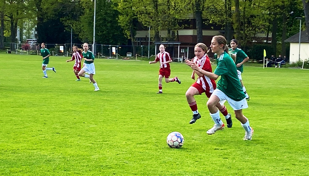 [{"clang_id":1,"value":"Fußballspiel: Spielerinnen in grünen und rot-weiß gestreiften Trikots auf grünem Rasenfeld. Eine Spielerin dribbelt den Ball. Im Hintergrund Bäume und Zuschauer."}]