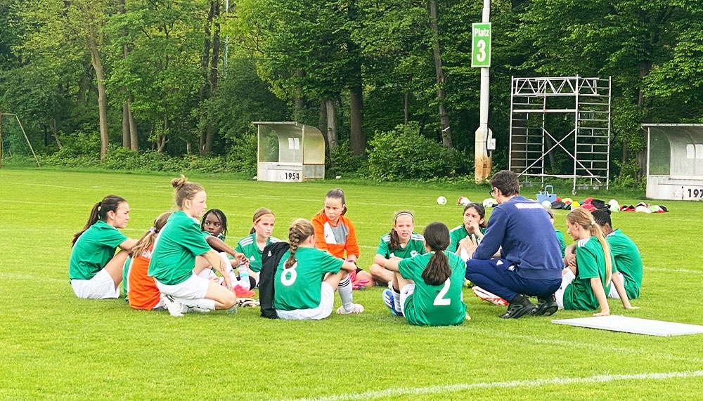 [{"clang_id":1,"value":"Fußballmannschaft im Kreis auf dem Rasen. Trainer erklärt Strategie. Im Hintergrund Bäume und Sportplatzgebäude."}]