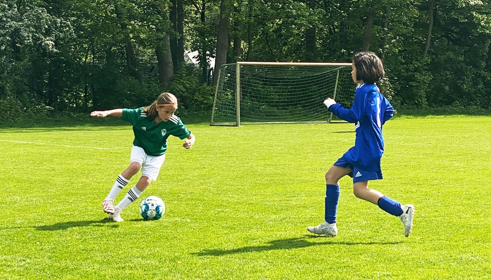 [{"clang_id":1,"value":"Zwei Kinder spielen Fußball auf einem grünen Rasenplatz. Links dribbelt ein Kind in grün-weißer Kleidung, rechts rennt ein Kind in blauem Trikot. Im Hintergrund ein Fußballtor und Bäume."}]