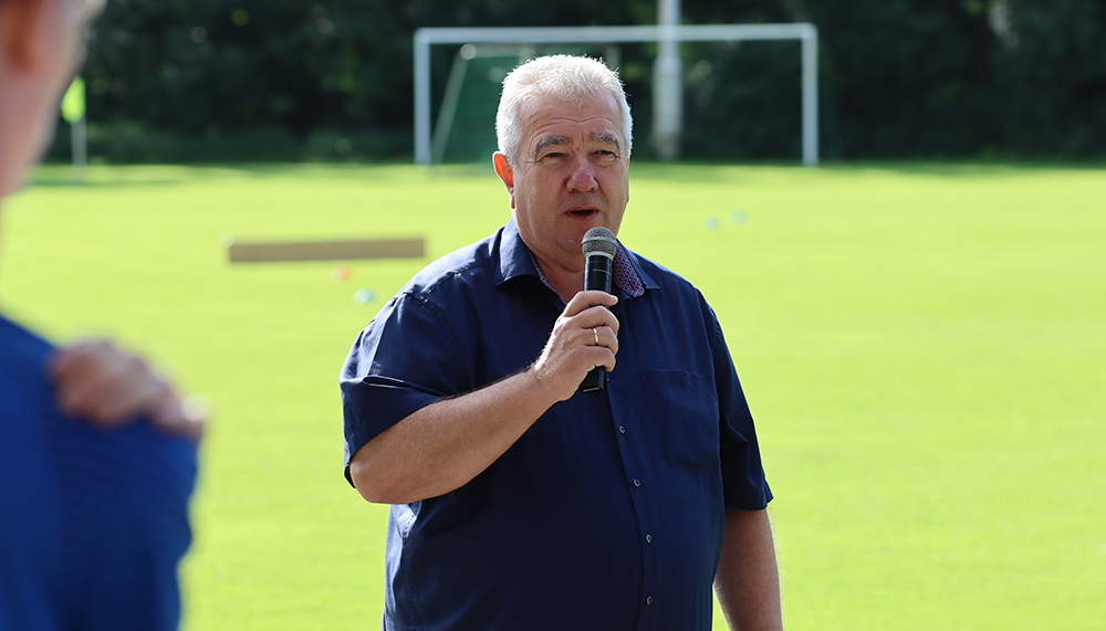 [{"clang_id":1,"value":"Mann mit Mikrofon steht auf Gras vor Fußballtor. Er trägt blaues Hemd und spricht. Im Hintergrund sind Bäume und Personen erkennbar."}]