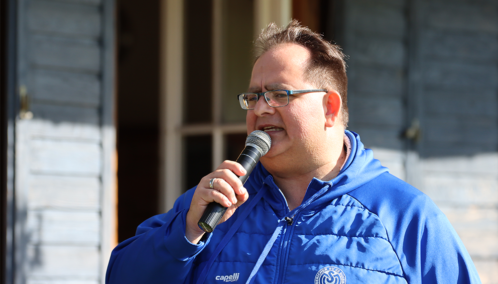 [{"clang_id":1,"value":"Mann mit Brille hält Mikrofon im Freien. Er trägt eine blaue Jacke mit Logo und spricht. Im Hintergrund ein Gebäude mit Holzverschalung."}]