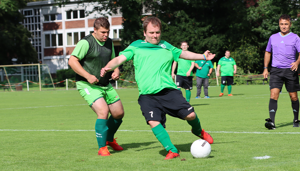 [{"clang_id":1,"value":"Fußballspiel: Zwei Spieler in grünen Trikots kämpfen um den Ball. Im Hintergrund weitere Spieler und ein Schiedsrichter. Grasplatz und Gebäude im Hintergrund."}]