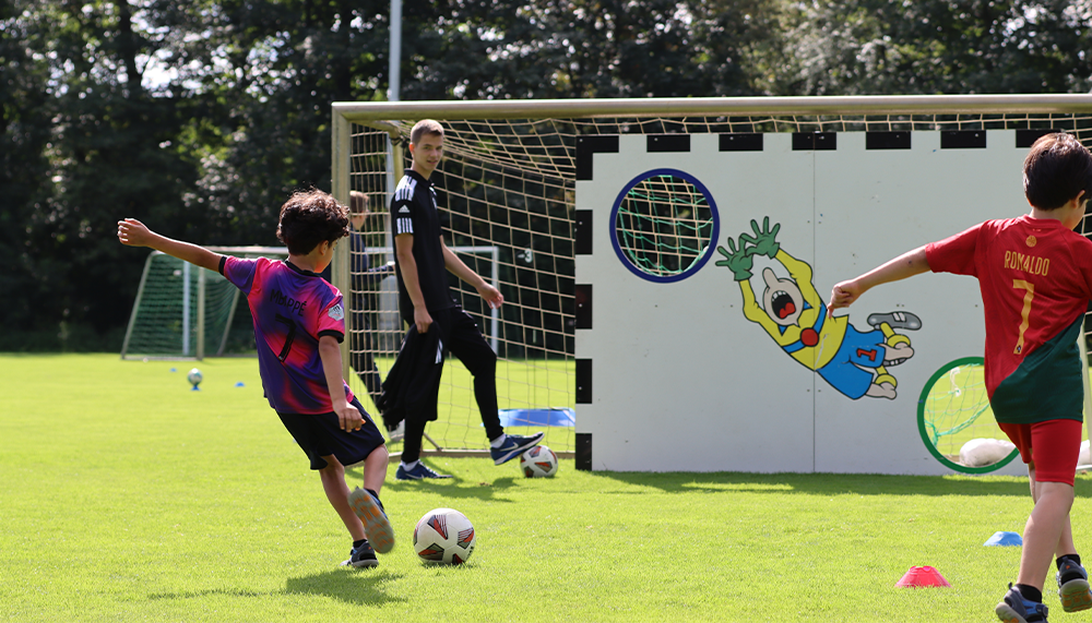 [{"clang_id":1,"value":"Zwei Kinder spielen Fußball auf einem grünen Rasen vor bunten Torwänden. Ein Trainer steht dahinter."}]