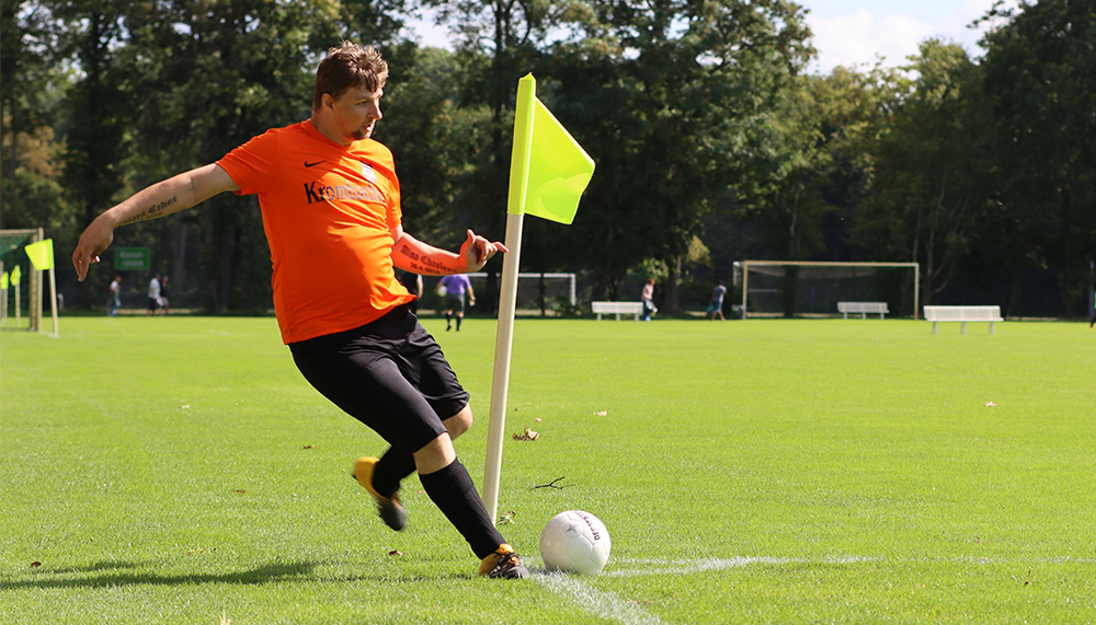 [{"clang_id":1,"value":"Fußballspieler in orangeem Trikot schießt Ball auf grüner Wiese am Eckfahnenmast. Im Hintergrund verschwommene Bäume und weitere Spieler."}]