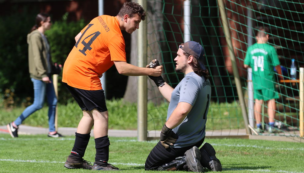 [{"clang_id":1,"value":"Fußballspiel: Spieler in orange und grau-schwarz helfen verletztem Torwart auf dem Rasen. Im Hintergrund weitere Spieler und das Tor."}]