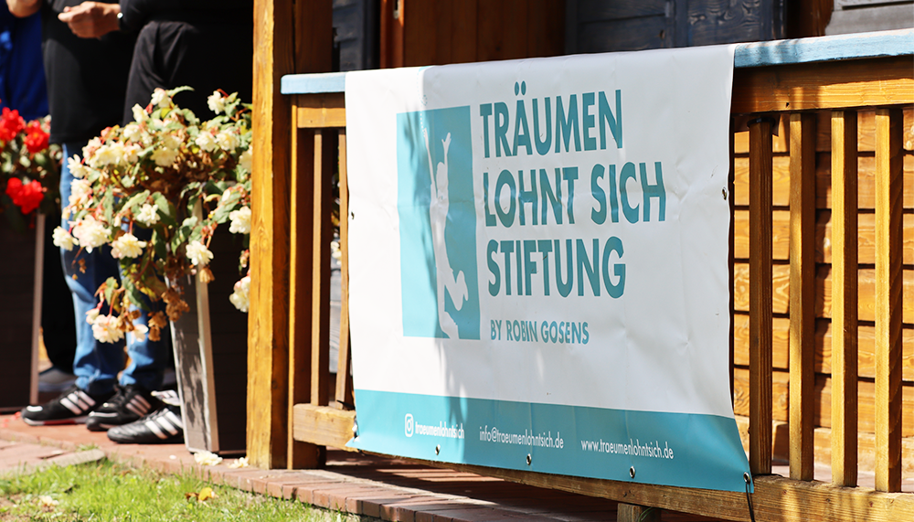 [{"clang_id":1,"value":"Banner \"Träumen lohnt sich Stiftung\" hängt an blauem Holzgeländer. Links Blumen, im Hintergrund Menschen und Gebäudeteile. Rasenfläche im Vordergrund."}]