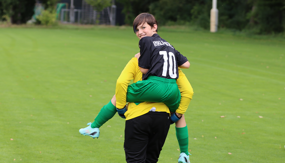 [{"clang_id":1,"value":"Zwei junge Fußballspieler auf einem Rasenfeld. Ein Spieler trägt den anderen auf seinen Schultern. Beide tragen Fußballtrikots und -shorts."}]