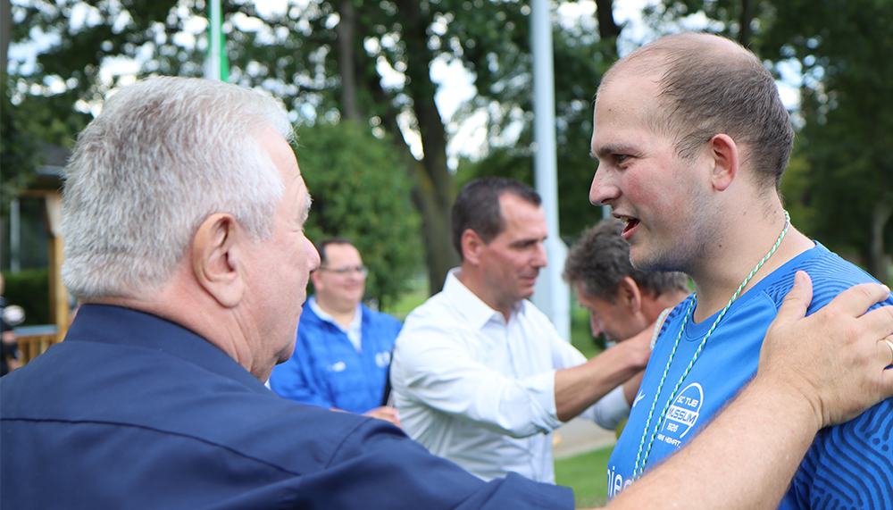 [{"clang_id":1,"value":"Zwei Männer unterhalten sich im Freien. Im Hintergrund weitere Personen und Bäume. Einer trägt Sportkleidung mit Werbeaufdruck."}]