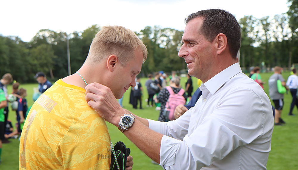 [{"clang_id":1,"value":"Zwei Männer stehen sich gegenüber. Der linke trägt ein gelbes Sporttrikot, der rechte ein weißes Hemd. Der Mann im Hemd legt dem anderen die Hand auf die Schulter und den Arm. Hintergrund: verschwommene Sportanlage und Personen."}]