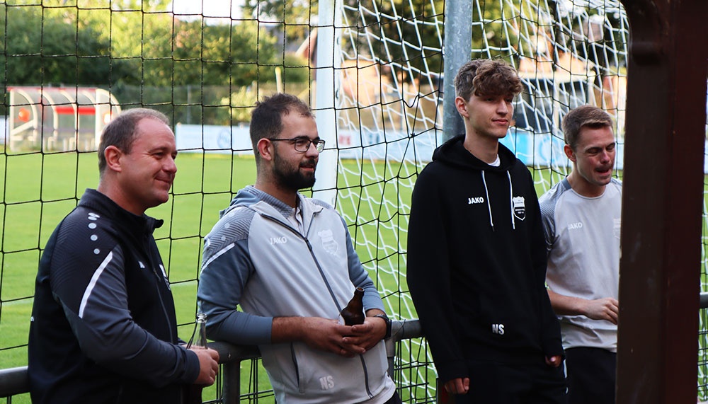[{"clang_id":1,"value":"Vier Männer stehen an einem Fußballfeld vor einem Tornetz. Links ein Mann mit dunklem Jackett, dann zwei Männer in grauen Trainingsanzügen und rechts ein jüngerer Mann mit schwarzem Hoodie."}]