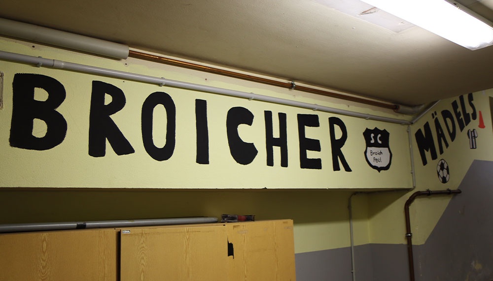 [{"clang_id":1,"value":"Wand mit großem, schwarzem Schriftzug \"BROICHER\" und \"MÄDELS\". Dazwischen ein Logo mit \"SC Broich\". Darunter Schränke. Beleuchtung durch Deckenlampen."}]