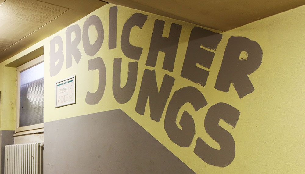 [{"clang_id":1,"value":"Gelbe Wand mit grauer Graffiti-Schrift \"BROICHER JUNGS\". Links ein Fenster und darunter ein Heizkörper. Schräge Schatten zeigen Licht von oben links."}]