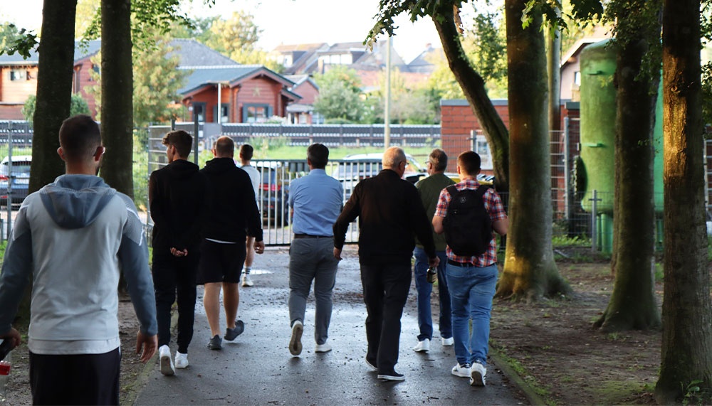 [{"clang_id":1,"value":"Eine Gruppe Männer geht auf einem Gehweg durch einen Park. Im Hintergrund sind Gebäude und Bäume zu sehen."}]
