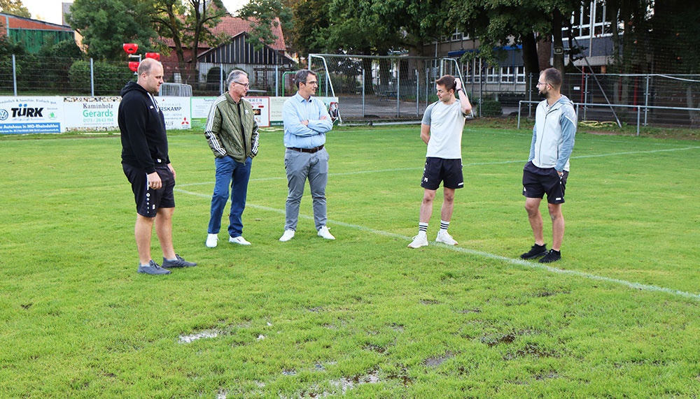 [{"clang_id":1,"value":"Fußballfeld mit sechs Männern, die in einer Reihe stehen und sich unterhalten. Im Hintergrund ein Zaun und Gebäude."}]