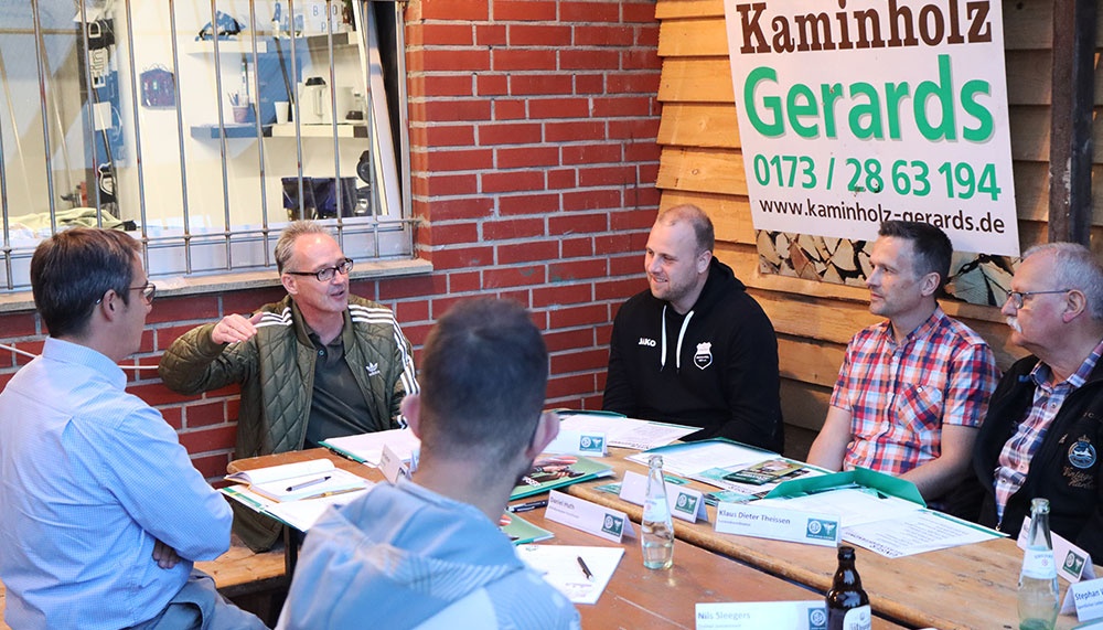 [{"clang_id":1,"value":"Sitzungsgruppe aus sechs Männern an einem Holztisch im Freien. Im Hintergrund ein Schild für Kaminholz Gerards und ein Schaufenster."}]
