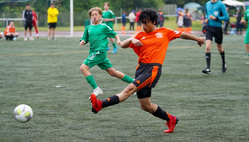 [{"clang_id":1,"value":"Zwei Fußballspieler im Spiel: Ein Spieler in Orange schießt auf das Tor, ein Gegenspieler in Grün versucht, den Schuss zu blocken. Im Hintergrund weitere Spieler und Zuschauer."}]