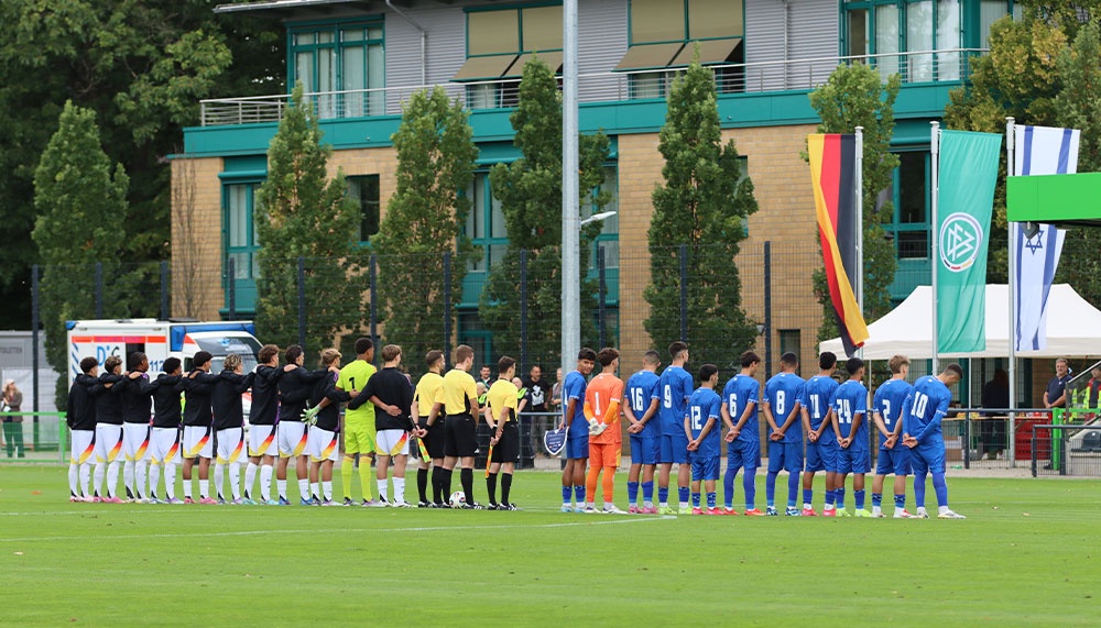 [{"clang_id":1,"value":"Fußballspiel: Zwei Mannschaften und Schiedsrichter stehen sich auf einem grünen Rasen gegenüber. Im Hintergrund ein Gebäude und Flaggen."}]