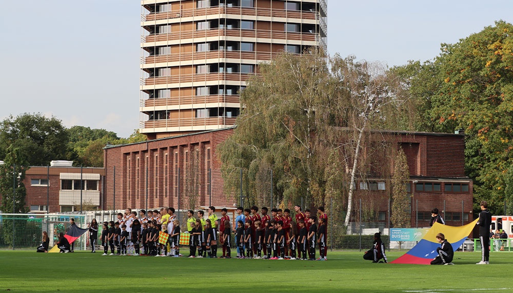Fußballfeld mit zwei Mannschaften in Trainingskleidung, umgeben von Bäumen und einem mehrstöckigen Backsteingebäude im Hintergrund.