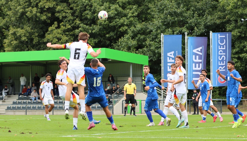 [{"clang_id":1,"value":"Fußballspiel: Zwei Spieler springen hoch, um einen Ball zu köpfen. Im Hintergrund weitere Spieler und Zuschauer auf einer Tribüne. Banner mit FIFA- und UEFA-Logo sind zu sehen."}]