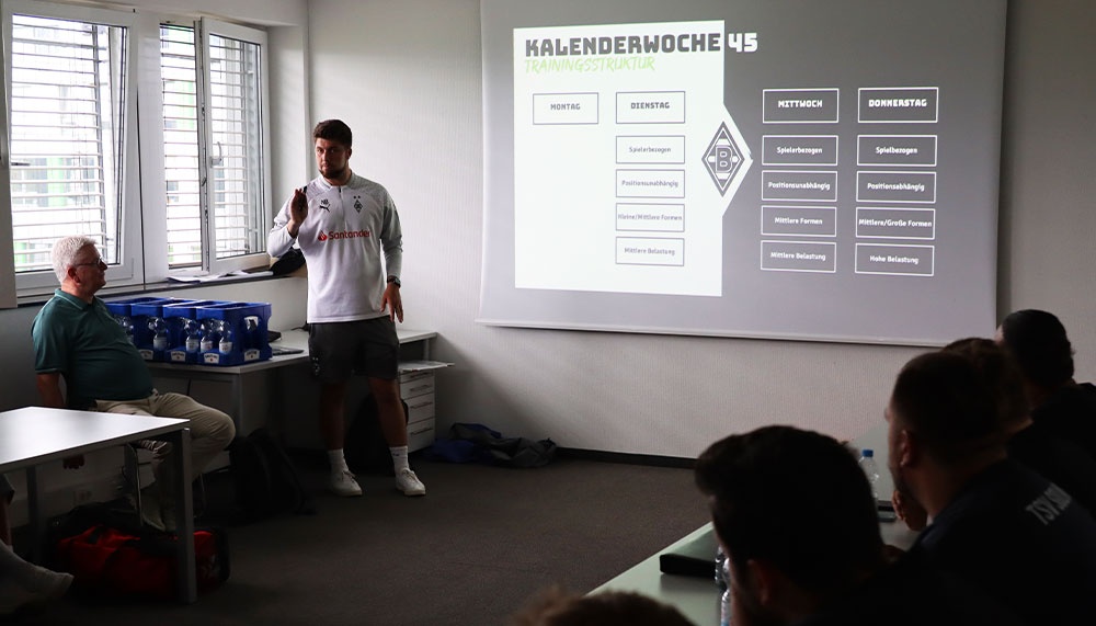 [{"clang_id":1,"value":"Trainer präsentiert Trainingsplan an Wand. Drei Personen sitzen an Tischen, beobachten die Präsentation. Heller Raum mit Fenster."}]