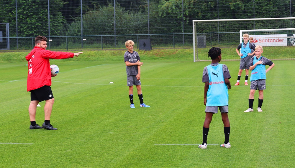 [{"clang_id":1,"value":"Fußballtrainer erklärt jungen Spielern im Trainingslager Anweisungen auf dem Rasen. Im Hintergrund ein Zaun und Fußballtor."}]