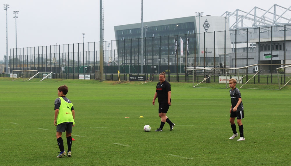 [{"clang_id":1,"value":"Drei Personen auf einem Fußballfeld. Links und rechts Kind in Sportkleidung, Mitte erwachsener Mann mit Fußball. Im Hintergrund Gebäude und Zaun."}]