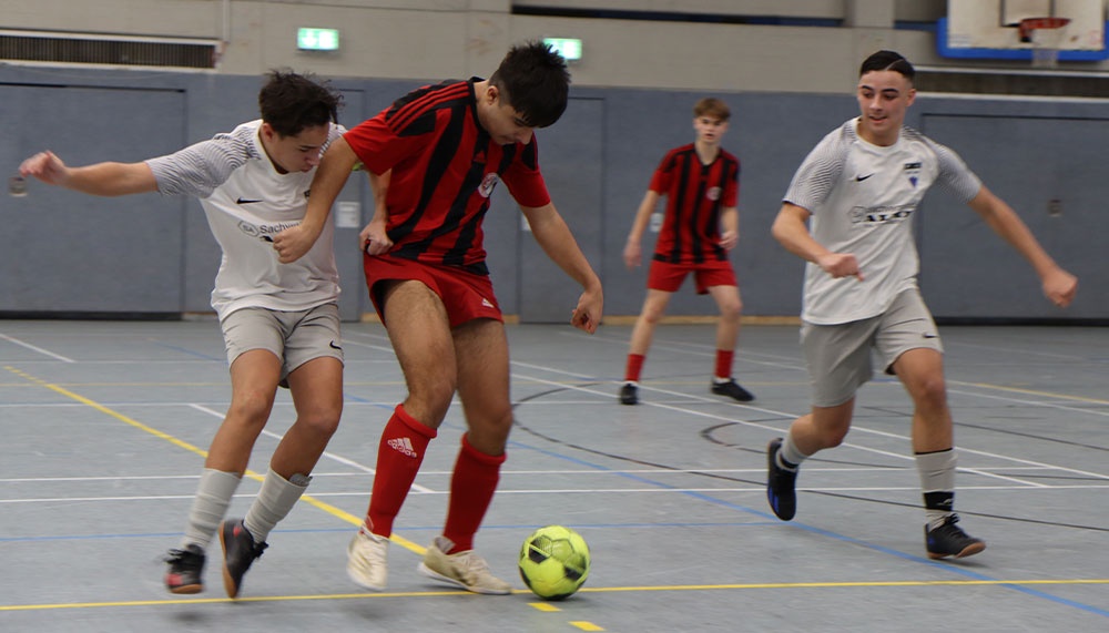 [{"clang_id":1,"value":"Futsal-Spiel in Sporthalle: Drei junge Männer in Sportkleidung spielen Fußball. Ein Spieler dribbelt den Ball, zwei weitere nähern sich."}]