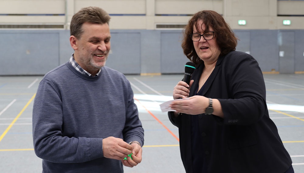 [{"clang_id":1,"value":"Zwei Personen in einer Sporthalle. Links ein Mann mit blauem Pullover, der lächelt. Rechts eine Frau mit Mikrofon, die spricht. Im Hintergrund eine Sporthalle."}]