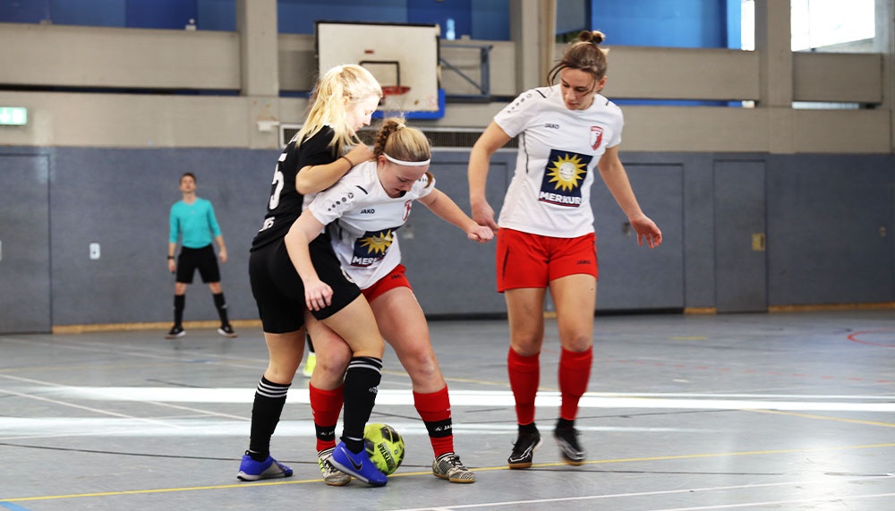 [{"clang_id":1,"value":"Drei Futsalspielerinnen in Sporthalle. Zwei in rot-schwarzer Ausrüstung kämpfen um den Ball, eine dritte in Schwarz blockt. Im Hintergrund weitere Personen."}]