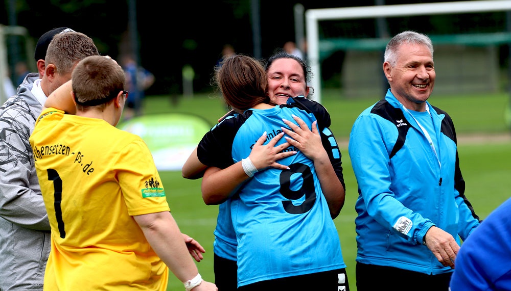 [{"clang_id":1,"value":"Fußballspieler feiern auf dem Rasen. Eine Spielerin in blauem Trikot wird von zwei Mitspielerinnen umarmt. Im Hintergrund weitere Personen und das Spielfeld."}]