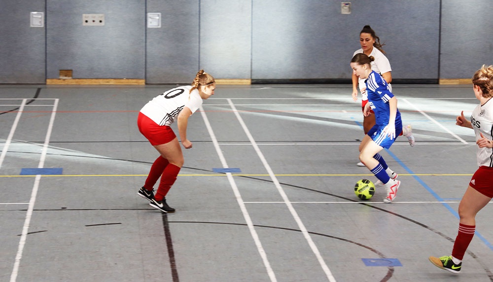 [{"clang_id":1,"value":"Drei Frauen spielen Hallenfußball. Eine Spielerin schießt auf den Ball, zwei weitere stehen in der Nähe. Sportlerinnen, Sport, Hallensport."}]
