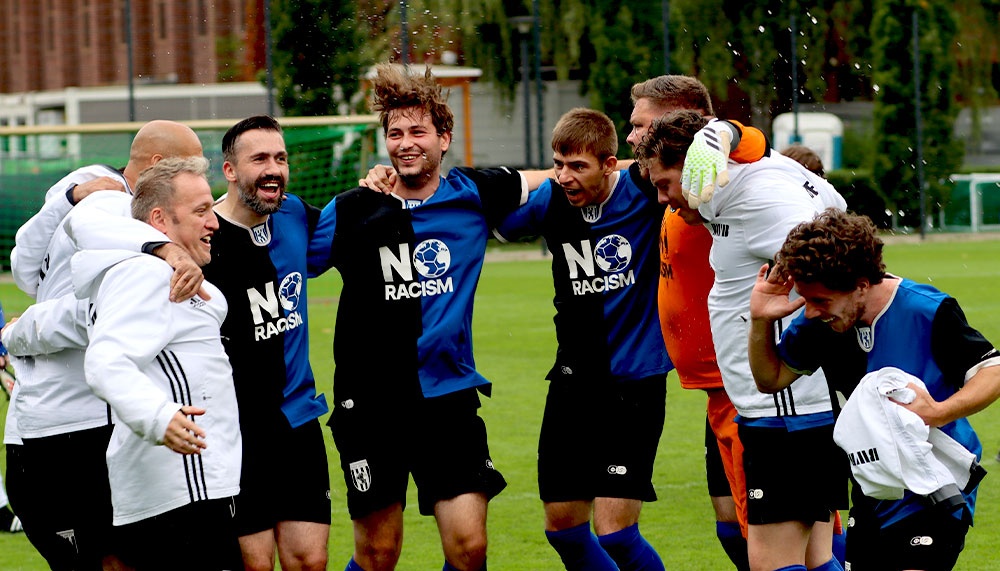 [{"clang_id":1,"value":"Fußballspieler feiern auf dem Rasen. Einige umarmen sich, andere stehen im Kreis. Sie tragen Trikots mit der Aufschrift \"NO RACISM\". Im Hintergrund ein Gebäude und Bäume."}]