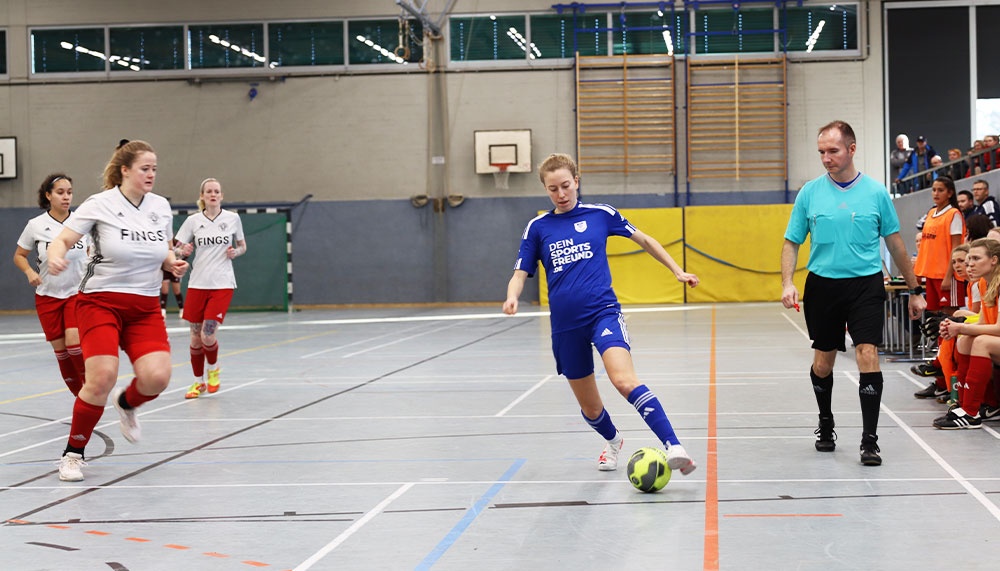 [{"clang_id":1,"value":"Fußballspiel in Sporthalle. Blaue Spielerin schießt Ball, während rote und orange gekleidete Gegnerinnen und ein Schiedsrichter in Schwarz-Weiß zusehen."}]