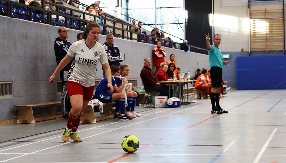 [{"clang_id":1,"value":"Futsalspiel in Sporthalle. Frau in weißem Trikot und roter Hose dribbelt Ball. Zuschauer auf blauen Tribünen im Hintergrund. Schiedsrichter mit erhobener Hand."}]