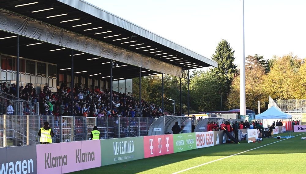[{"clang_id":1,"value":"Fußballstadion mit Zuschauern unter einem überdachten Tribünenbereich. Im Vordergrund Werbebanner, dahinter grüner Rasen und im Hintergrund Bäume."}]