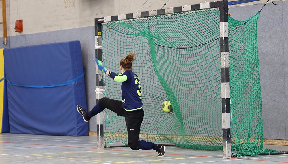[{"clang_id":1,"value":"Handballtorwartin in blauer und schwarzer Sportkleidung springt zur Ballabwehr. Im Hintergrund blaue Wandpolsterung und ein Handballfeld."}]