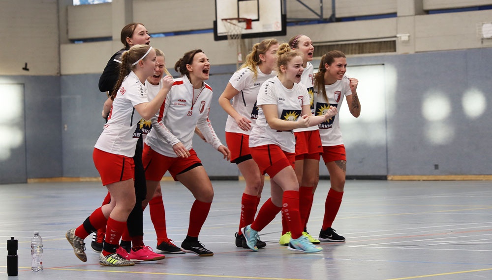 [{"clang_id":1,"value":"Handballteam jubelt eng umschlungen in Sporthalle. Spielerinnen tragen rote und weiße Trikots und Shorts. Im Hintergrund Basketballkorb und Zuschauer."}]