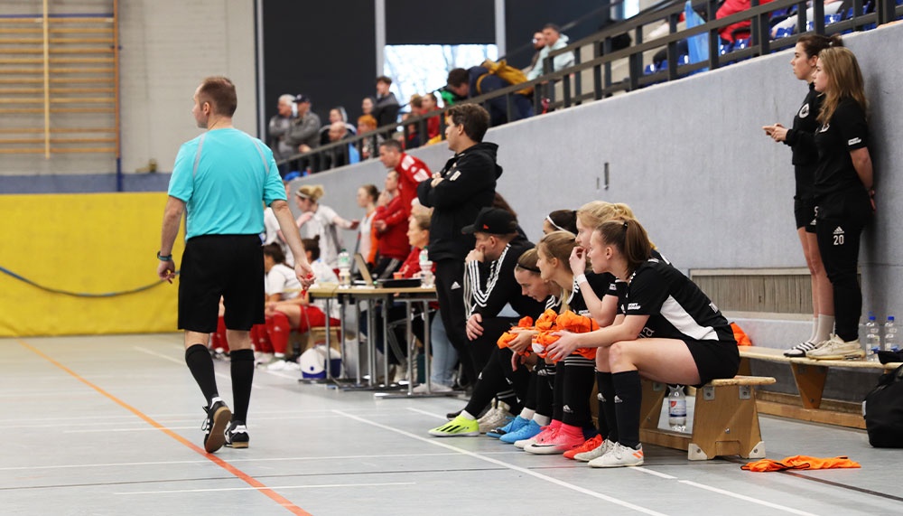 [{"clang_id":1,"value":"Handballspiel: Schiedsrichter mit zurückgekehrtem Oberkörper, Spielerinnen auf der Bank, Zuschauer auf Tribüne. Sporthalle mit hellen Wänden und roter Markierung."}]