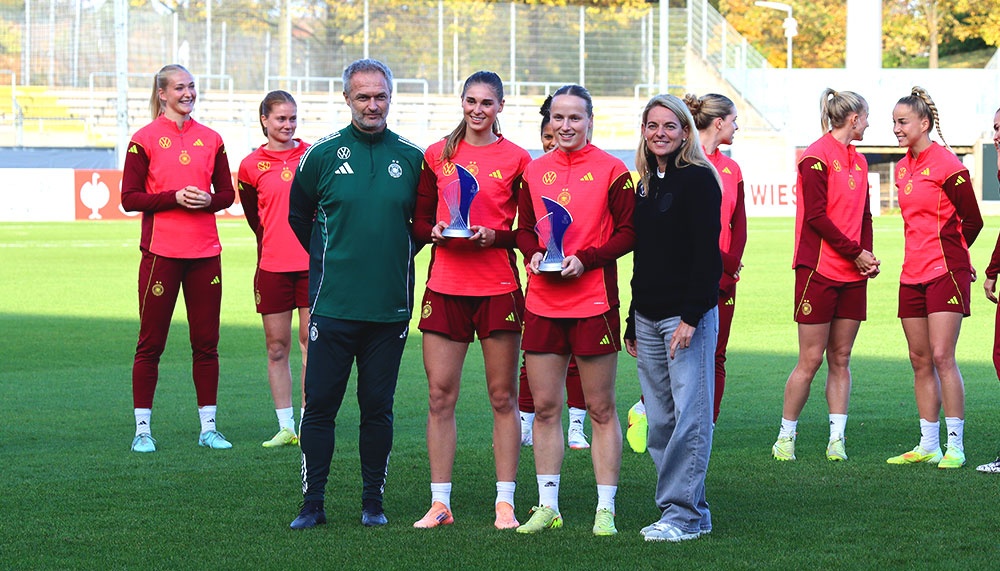 [{"clang_id":1,"value":"Fußballspielerinnen in roten Trainingsanzügen mit Trainer und Frau, die Trophäen halten. Im Hintergrund ein Fußballfeld und Gebäude."}]