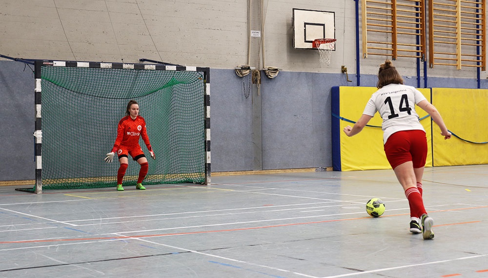 [{"clang_id":1,"value":"Futsalspiel in Sporthalle: Spielerin mit rotem Trikot schießt auf Tor, Torwartin im Gegenstand. Basketballkorb im Hintergrund."}]