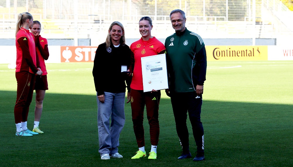 [{"clang_id":1,"value":"Drei Frauen und ein Mann stehen auf einem Fußballfeld. Die Frau in der Mitte hält eine Urkunde. Alle tragen Sportkleidung mit dem DFB-Logo. Im Hintergrund weitere Sportlerinnen."}]